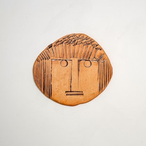 May include: A flat, circular terracotta-colored ceramic plaque featuring an incised, stylized human face. The design includes etched lines for hair, two circular eyes, a straight nose line, and a horizontal line for the mouth.