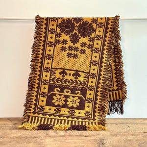 Peut inclure: Une couverture tissée en brun et or avec un motif géométrique. La couverture a des franges sur tous les côtés.