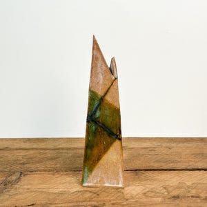 Puede incluir: Un jarrón de cerámica alto y angular con un diseño geométrico. El jarrón presenta una forma triangular con una base marrón, acentuada por secciones verdes y marrones. Líneas negras definen el patrón geométrico. El jarrón se exhibe sobre una superficie de madera.