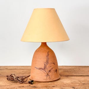 Puede incluir: Una lámpara de mesa de color terracota con una pantalla beige. La base de la lámpara tiene forma de campana y está decorada con un diseño floral marrón. La lámpara está sobre una superficie de madera.