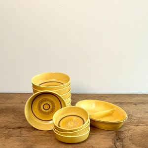 Peut inclure: Un ensemble de bols en céramique jaune avec un motif géométrique brun. Les bols sont empilés en pile, avec un grand bol à l'avant. Il y a aussi un plat divisé en céramique jaune avec un bord brun.
