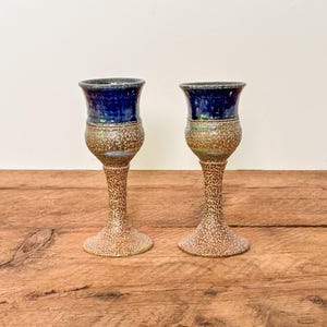 Puede incluir: Dos cálices de cerámica con un borde esmaltado en azul profundo y una base texturizada moteada en tonos beige y marrón. Cada recipiente tiene una abertura acampanada, un cuerpo redondeado y un tallo delgado que termina en una base circular estable.