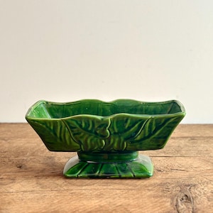 May include: A green ceramic planter with a leaf design. The planter has a square base and a rectangular top.
