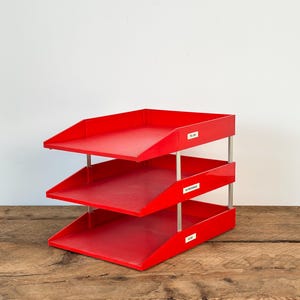 May include: A red three-tier desk organiser with silver support rods. Each tray has a label with text. The organiser sits on a wooden surface against a white background. The labels read "TO DO", "IN PROGRESS", and "DONE".