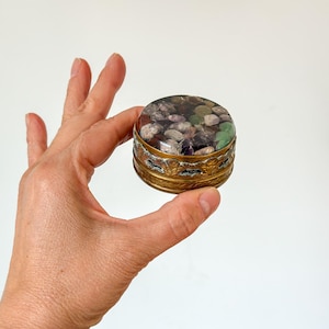 Puede incluir: Una pequeña caja redonda para baratijas con una base de color latón y una tapa llena de piedras de colores. Las piedras son una mezcla de morado, verde y blanco. La caja se sostiene en una mano sobre un fondo blanco.