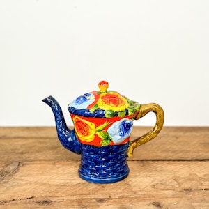 May include: A hand-painted ceramic teapot with a blue woven basket design base. The lid and upper portion feature a floral pattern with red, yellow, and blue roses. The handle is a golden brown colour.
