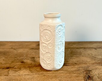 May include: A white ceramic vase with a textured surface featuring embossed spiral patterns. The vase has a cylindrical body with a slightly tapered neck and a smooth rim, showcasing a consistent matte finish across its entire exterior structure.