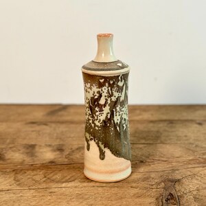 May include: A small, white ceramic vase with a brown and white speckled glaze. The vase has a rounded bottom and a narrow neck.