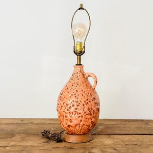 May include: A vintage ceramic table lamp with a textured, speckled orange glaze. The lamp has a jug-like shape with a handle, a brass neck, and a bulb. The lamp sits on a wooden base and has a visible power cord.