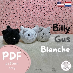 Puede incluir: Tres juguetes de gato de ganchillo en negro, azul claro y gris. Los juguetes se exhiben sobre una superficie blanca y suave contra un fondo rosa con pequeños patrones de corazón. El texto "Billy Gus Blanche" es visible.
