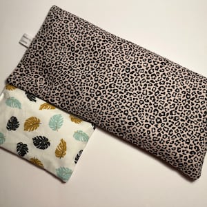 Op de afbeelding: Twee stoffen oogkussens, een met een roze achtergrond en een zwarte luipaardprint, en de andere met een witte achtergrond en een blauw en bruin bladmotief. Het kussen met de luipaardprint heeft een label met de tekst "Handmade by Leonie".