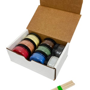 May include: A white cardboard box with the lid open, revealing six small jars of colorful powder. The jars are stacked in a row, with the colors red, orange, yellow, green, blue, and white. A bundle of wooden sticks is also included.