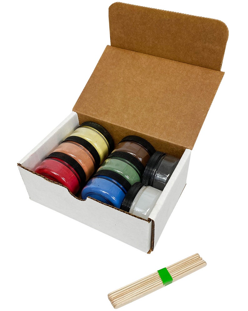 May include: A white cardboard box with the lid open, revealing six small jars of colorful powder. The jars are stacked in a row, with the colors red, orange, yellow, green, blue, and white. A bundle of wooden sticks is also included.