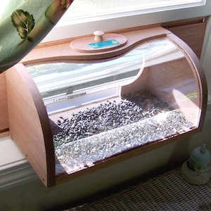 May include: A wooden window bird feeder with a clear curved front, filled with birdseed. The feeder has a wooden lid with a small handle. The bird feeder is mounted on a window sill.