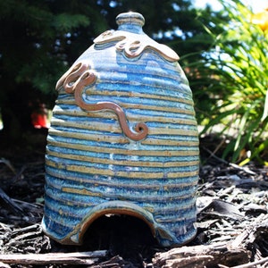 Op de afbeelding: Een handgemaakte keramische bijenkorf met een blauw en bruin gestreept patroon. De bijenkorf heeft een bruine werveldecoratie en een ronde opening aan de basis. De bovenkant heeft een decoratief wervelontwerp.