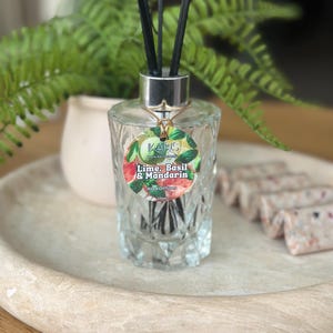 May include: A clear glass reed diffuser with black reeds, labelled "Lime, Basil & Mandarin". The diffuser sits on a wooden tray with a small white pot and green plant in the background. The diffuser is 170ml.