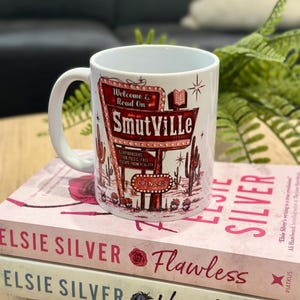 May include: A white ceramic mug with a retro sign design that reads "SmutVille", with cacti and the words "Welcome & Read On." The mug is placed on top of two books by Elsie Silver, titled "Flawless" and "Heartless."