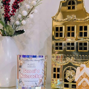 May include: A clear glass candle with the text "Son of a Nutcracker" and a gold-coloured house-shaped decoration. The candle has a label with the words "Ginger, Nutmeg, Cinnamon" and the brand name "KARIS". A white vase with red berries and white balls is also in the image.
