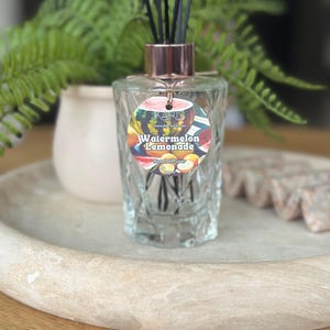May include: A clear glass reed diffuser with black reeds and a rose gold collar. The label reads "Watermelon Lemonade" with a colourful watermelon design. The diffuser sits on a light-coloured wooden tray.