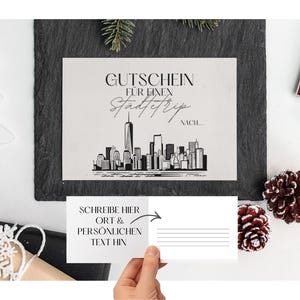 Pode incluir: Um vale-presente com uma ilustração do horizonte de Nova York. O texto diz "Gutschein für einen Städtetrip nach..." e "Schreibe hier Ort & persönlichen Text hin". A imagem está em uma ardósia preta com elementos decorativos.