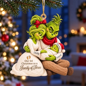 May include: A hanging Christmas ornament featuring a green family of three characters. The female figure wears a white dress with the text 'Emily' and 'Our 1st Christmas as a Family of Three'. The male figure wears a red scarf, and the baby wears a red hat with 'Thomas' and 'David'.