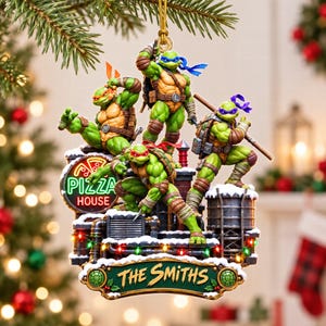 May include: A decorative hanging ornament featuring four green Teenage Mutant Ninja Turtles characters posed on a snowy rooftop with a neon Pizza House sign and a green plaque at the bottom with the text The Smiths in gold lettering.