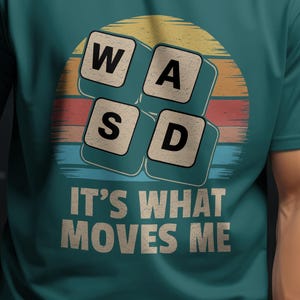以下が含まれることがあります： キーボードのキーを表すW、A、S、Dの文字がレトロな夕焼けのグラフィックで描かれたターコイズブルーのTシャツ。グラフィックの下に「IT'S WHAT MOVES ME」というテキストが印刷されています。
