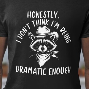 Puede incluir: Camiseta negra con un gráfico blanco de un mapache con un sombrero de vaquero y un pañuelo. El texto "Honestamente, no creo que esté siendo lo suficientemente dramático" está impreso debajo del mapache.