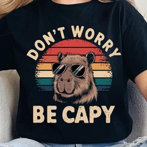 Puede incluir: Una camiseta negra con un gráfico vintage de un capibara con gafas de sol. El gráfico dice "Don't Worry Be Capy".