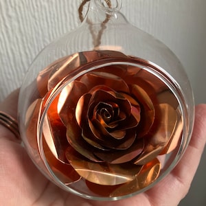 May include: A copper rose encased in a clear glass globe ornament. The rose is made of thin copper sheets and has a delicate, layered appearance.