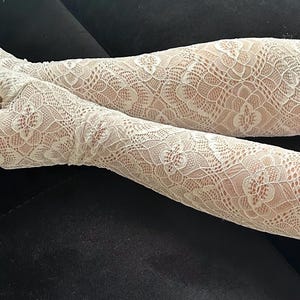 Puede incluir: Calentadores de brazo de encaje blanco con un patr&oacute;n floral. Estos guantes sin dedos se extienden desde la mu&ntilde;eca hasta la parte superior del brazo. El delicado dise&ntilde;o de encaje es visible sobre la piel. Los calentadores de brazo son de color claro y neutro.