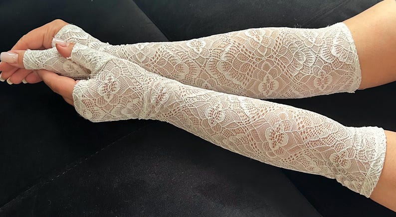 Puede incluir: Calentadores de brazo de encaje blanco con un patr&oacute;n floral. Estos guantes sin dedos se extienden desde la mu&ntilde;eca hasta la parte superior del brazo. El delicado dise&ntilde;o de encaje es visible sobre la piel. Los calentadores de brazo son de color claro y neutro.