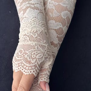 Op de afbeelding: Ivoor kanten vingerloze handschoenen met ingewikkelde bloempatronen. De handschoenen zijn gemaakt van delicate, transparante kant en zijn ontworpen om de hand en onderarm te bedekken, waarbij de vingers vrij blijven.
