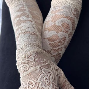 Op de afbeelding: Ivoorwitte kanten handschoenen met een delicaat bloemmotief. De handschoenen zijn vingerloos en reiken tot aan de arm. Het ingewikkelde ontwerp en de lichte kleur cre&euml;ren een elegante, vintage uitstraling, perfect voor speciale gelegenheden.