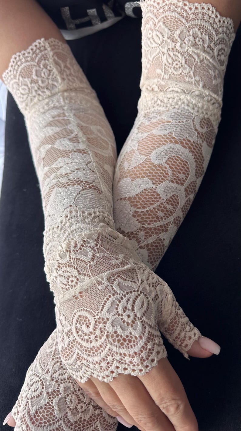 Op de afbeelding: Ivoorwitte kanten handschoenen met een delicaat bloemmotief. De handschoenen zijn vingerloos en reiken tot aan de arm. Het ingewikkelde ontwerp en de lichte kleur cre&euml;ren een elegante, vintage uitstraling, perfect voor speciale gelegenheden.