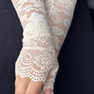 Op de afbeelding: Ivoor kanten vingerloze handschoenen. De handschoenen hebben een ingewikkeld bloemenpatroon en zijn gemaakt van delicate, transparante kant. De handschoenen zijn ontworpen om de hand tot aan de pols te bedekken, met een open ontwerp voor de vingers.