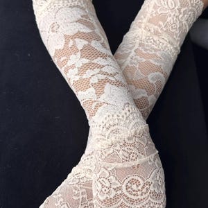 Op de afbeelding: Ivoor kleurige kanten handschoenen met een bloemenpatroon. De handschoenen zijn vingerloos en reiken tot aan de arm. Het kant is gedetailleerd en delicaat, met een geschulpte rand bij de pols. De achtergrond is zwart.