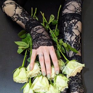 May include: A bouquet of white roses with green stems, held by a person wearing black lace gloves. The person is wearing a black strapless top and has a butterfly tattoo on their shoulder.