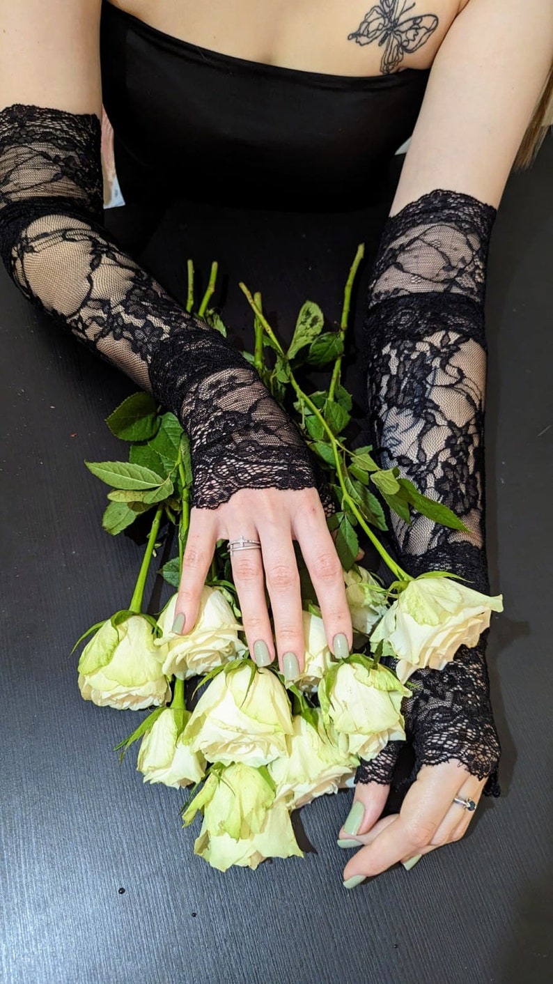 May include: A bouquet of white roses with green stems, held by a person wearing black lace gloves. The person is wearing a black strapless top and has a butterfly tattoo on their shoulder.