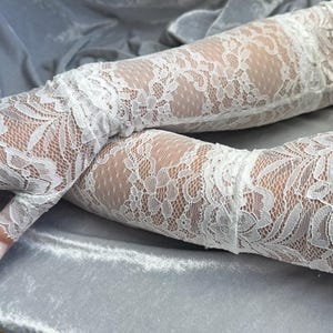 Puede incluir: Un par de guantes de encaje blanco con un patrón floral. Los guantes son largos y llegan más allá de los codos.