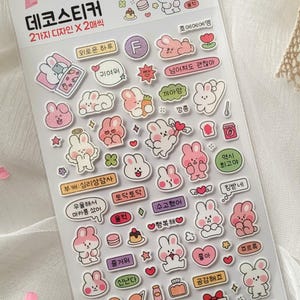 Peut inclure: Une planche d'autocollants décoratifs avec divers lapins de dessins animés roses, blancs et autres couleurs pastel. Les autocollants comprennent des bulles de texte et de petites illustrations de nourriture et de cadeaux. La planche est emballée dans une pochette en plastique transparent.