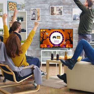May include: Four friends are gathered around a television watching a Halloween-themed game show called "Game of Thrones". The television screen is decorated with pumpkins, bats, and a spooky forest. The friends are all smiling and cheering.