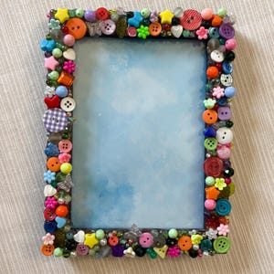 May include: A rectangular picture frame decorated with a dense arrangement of colorful plastic buttons, beads, and small charms in various shapes including stars, flowers, and butterflies, surrounding a light blue watercolor-style background.