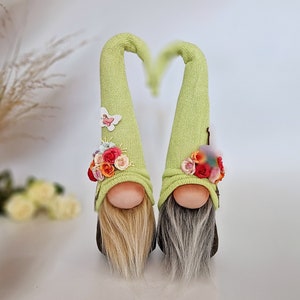 May include: Two gnomes with green knit hats decorated with flowers and butterflies. The gnomes have long white and blonde beards.