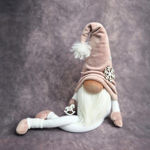 Könnte beinhalten: Ein plüschiger rosa Zwerg mit weißem Bart und einem weißen Fellpompon auf seiner Mütze. Der Zwerg hält ein kleines Holz-Schaukelpferd und hat einen Holz-Schneeflocken-Anhänger auf seiner Mütze.