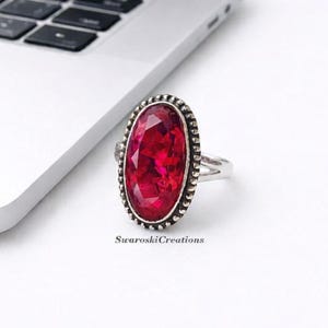 May include: A silver ring with a large, oval, ruby-red gemstone. The gemstone is surrounded by a beaded border. The ring is on a white surface next to a laptop. The text "SwaroskiCreations" is below the ring.