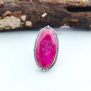 May include: A silver ring featuring a large, oval-shaped, vibrant pink gemstone. The gemstone is set in a silver band with a twisted rope design around the edge. The ring is displayed on a white surface with a piece of wood in the background.