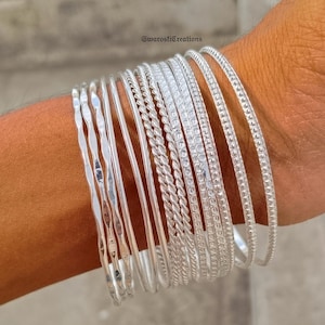 May include: A selection of silver bangles in diverse designs, including hammered, twisted, and beaded textures. The bangles are stacked on a wrist, displaying their different styles and reflective surfaces. The image is well-lit, highlighting the metallic shine of the jewellery.