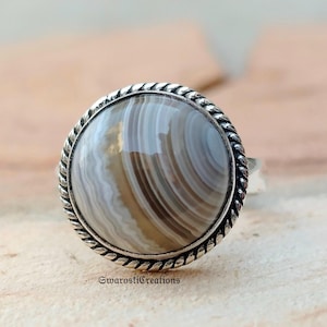 Puede incluir: Un anillo de plata con una piedra grande, redonda y pulida. La piedra presenta un patrón arremolinado de bandas grises y beige. El bisel del anillo tiene un diseño de cuerda trenzada. El texto "SwarostiCreations" está en la parte inferior del anillo.