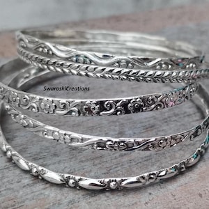 May include: Set of five silver bangles with intricate floral and geometric designs. The bangles are stacked on top of each other, creating a layered look.